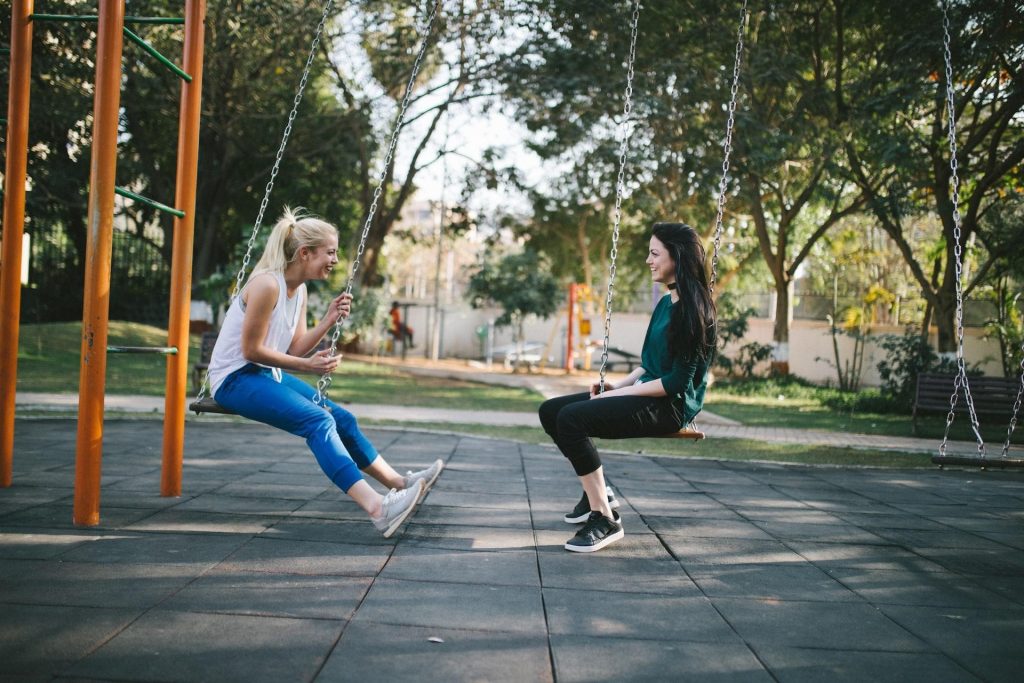 Kaksi nuorta naista istuu vastakkain leikkipuiston keinuissa. Naiset hymyilevät toisilleen.