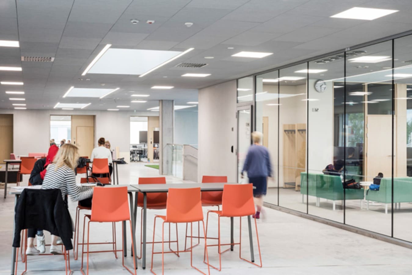 Valoisa kampustila, pöytiä joiden äärellä istuu ihmisiä, ohitse kävelee ihminen takaa päin kuvattuna.