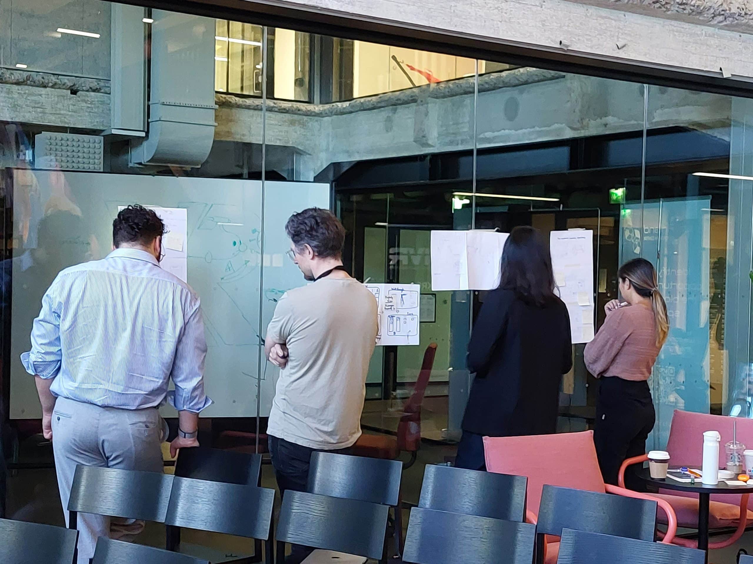 Four people facing a glass wall, their backs turned to camera. On the wall, sheets of paper and notes. In front of photo, empty seats.