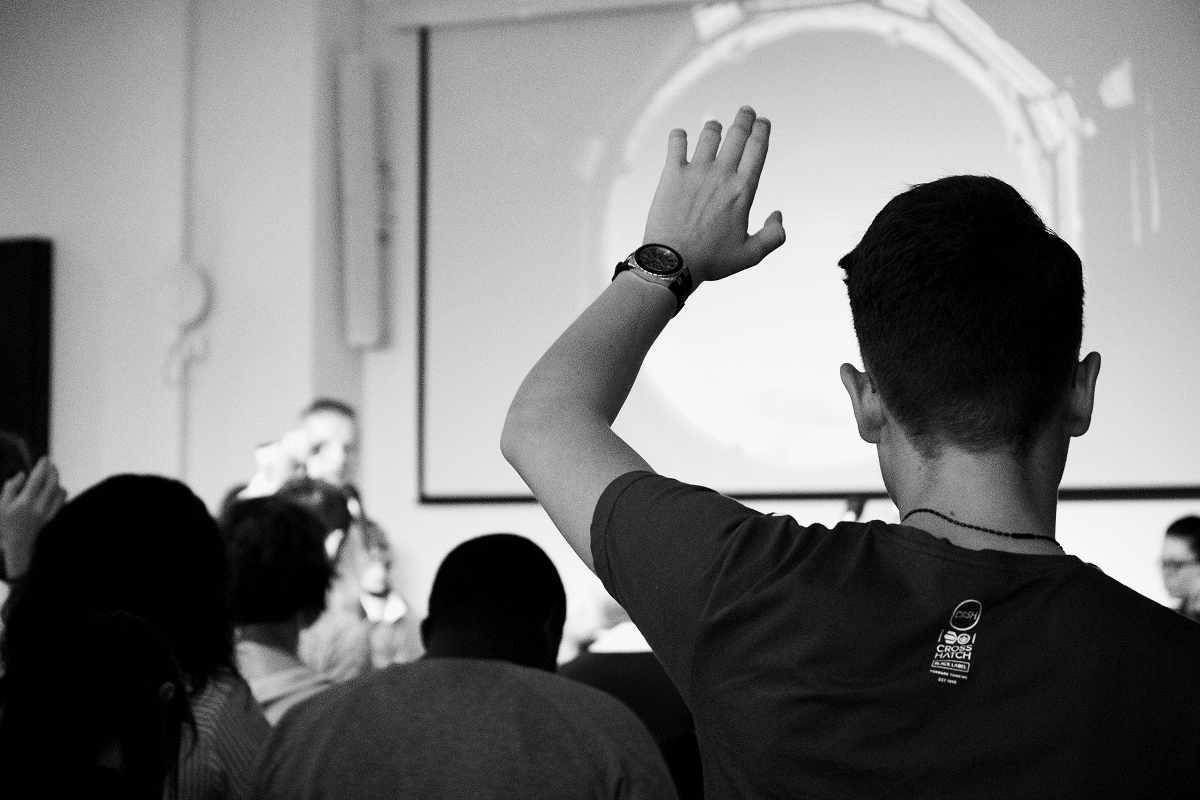 Black and white photo of audience from the back, one hand raised, speaker on stage facing the camera
