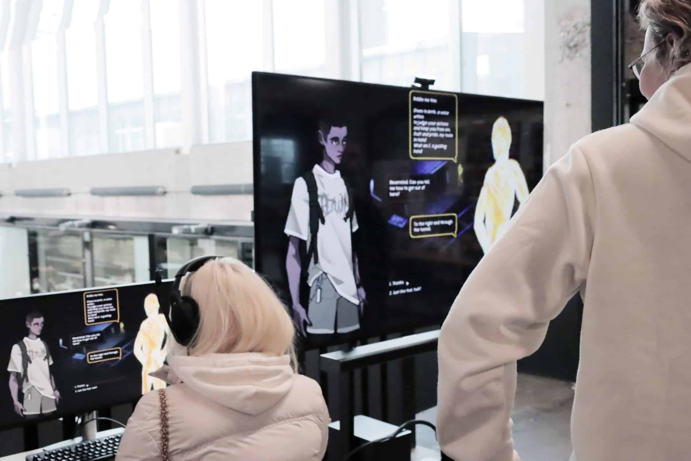 Screens at game design studio, two people back to camera, one standin gon right edge and other seated on left corner.