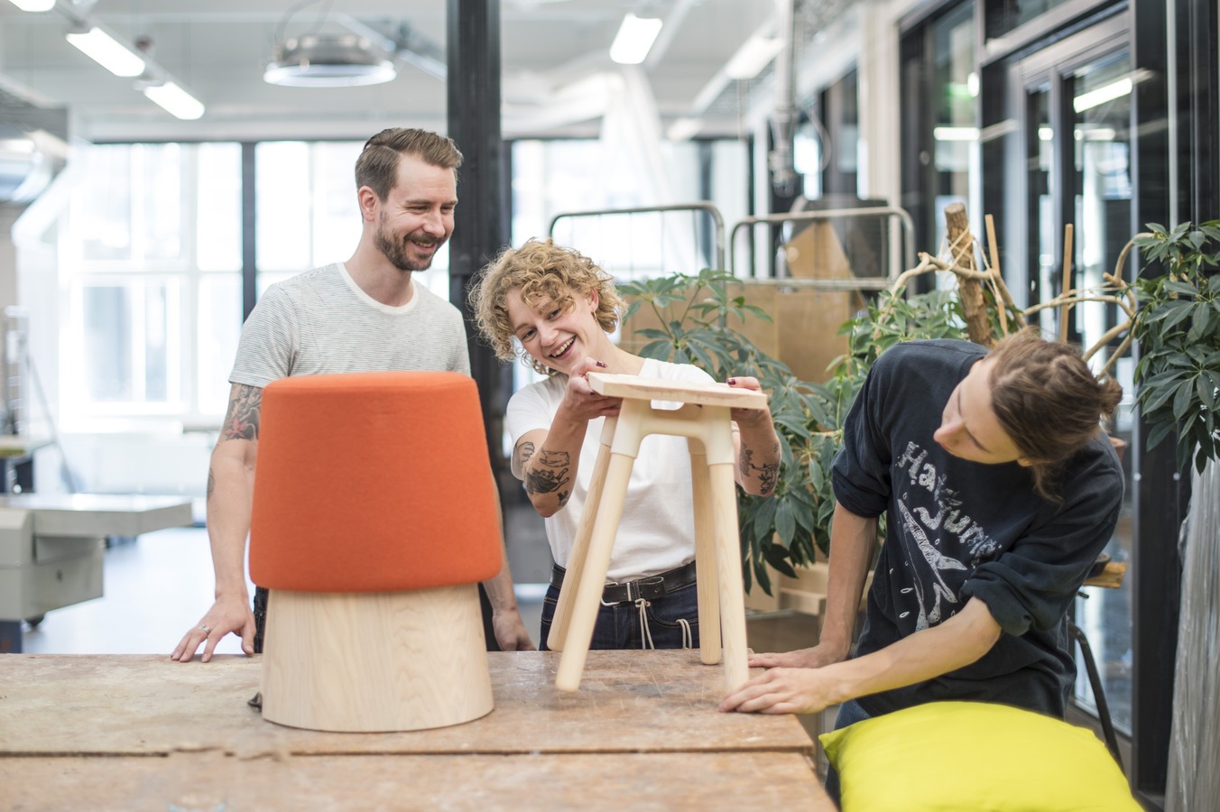 Lasiseinäisessä huoneessa pöytä, pöydän päällä jakkaroita, pöydän ympärillä kolme henkilöä tarkastelemassa yhden jakkaran ominaisuuksia.